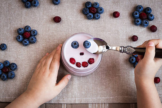 A child's hand enjoying yogurt topped with blueberries and raspberries on a textured tablecloth.