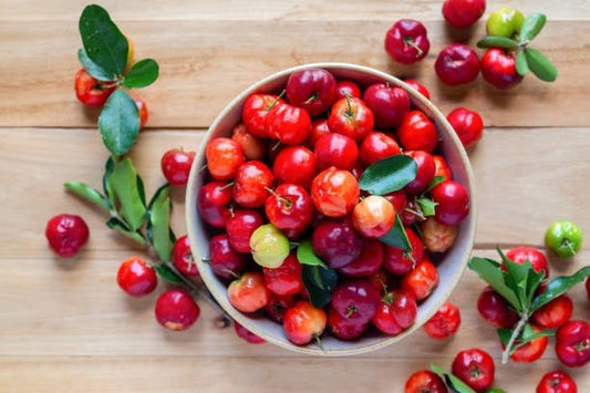 Bowl of fresh acerola cherries surrounded by leafy branches on a wooden surface.