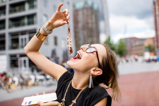 Woman joyfully enjoying a treat outdoors, holding a piece of food with glasses and a stylish outfit.