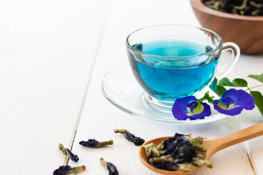 A cup of blue butterfly pea tea with dried flowers and fresh blue petals on a wooden spoon.