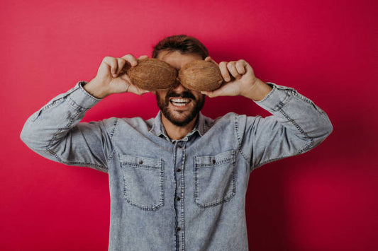 Man holding coconuts in front of his face, smiling against a vibrant pink background.