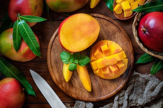 Freshly cut mango slices on a wooden board surrounded by whole mangos and green leaves.