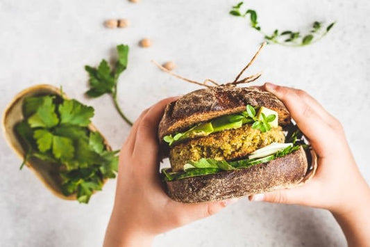 Hands holding a fresh veggie burger stacked with greens, served on a rustic table with herbs.