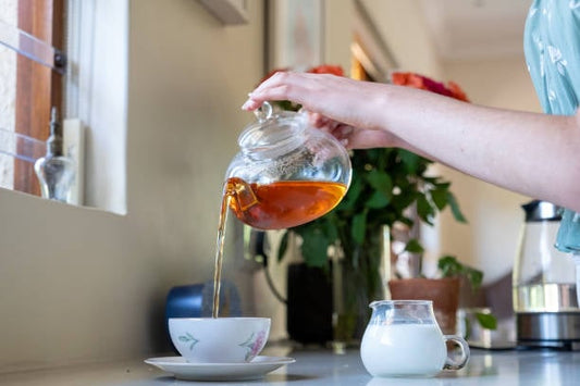 A hand pouring rooibos tea from a glass teapot into a white cup on a kitchen counter.