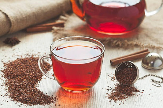A glass cup of red rooibos tea surrounded by loose tea leaves and a tea infuser.