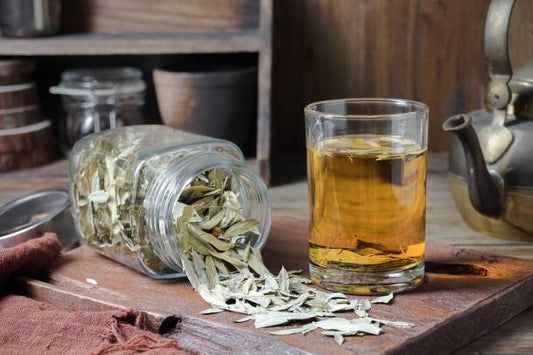 Jar of senna leaves spilling onto a wooden surface next to a glass of brewed tea.
