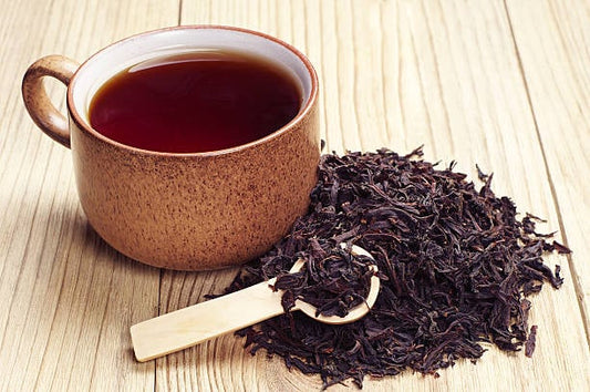 A cup of black tea with loose leaf tea beside a wooden spoon on a wooden surface.
