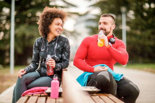 Two athletes enjoying sports drinks together after a workout, seated on a bench outdoors.