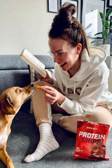 Woman enjoying BioTechUSA paprika protein chips with her dog while reading a book at home.