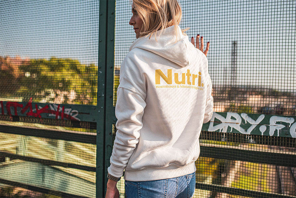 Person wearing a Nutri hoodie, standing on a bridge with cityscape in the background and graffiti on the fence.