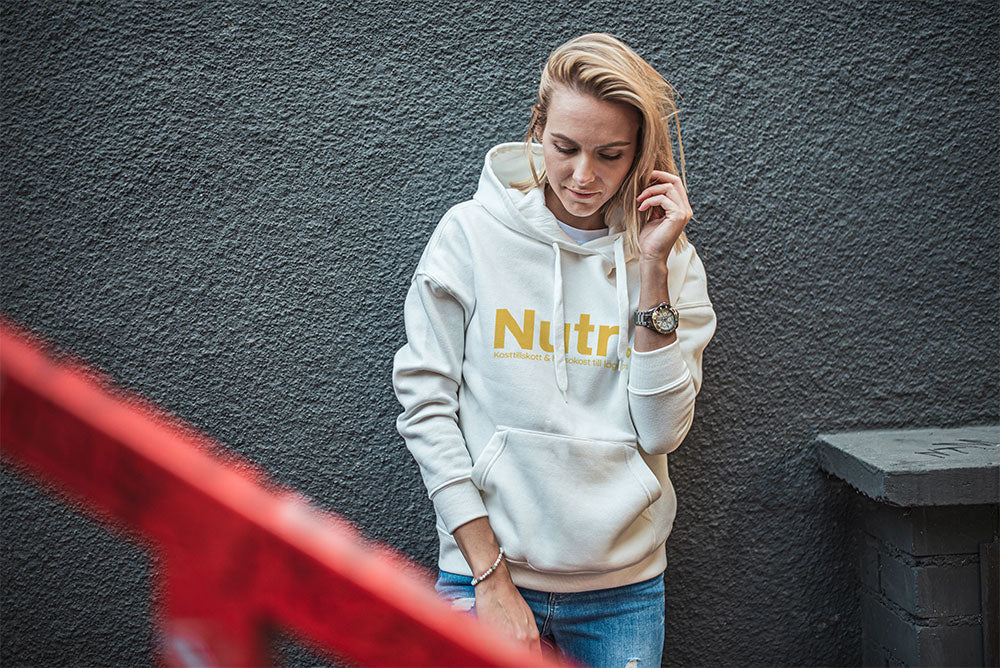 Woman in a white hoodie with 'Nutri' logo, standing against a textured gray wall, showcasing casual streetwear style.