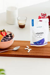 Nutriversum D3 + K2 vitamin capsules beside a bowl of fruit and a cup of coffee on a wooden table.