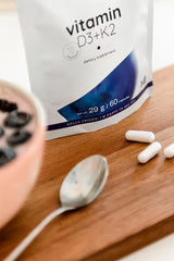 Nutriversum D3 + K2 vitamin dietary supplement with capsules and smoothie bowl on a wooden surface.