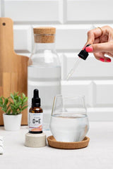 Hand adding Nutriversum Vitamin D+E drops to a glass of water on a kitchen counter.