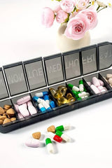 Oly Maxi Pill Box with seven compartments filled with various pills and supplements, beside a vase of pink roses.