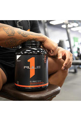 Rule One R1 Protein container on gym bench, showcasing high-quality whey isolate for muscle recovery.