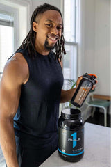 Smiling man holding Rule One R1 Whey Blend protein powder and shaker in kitchen, showcasing fitness lifestyle.