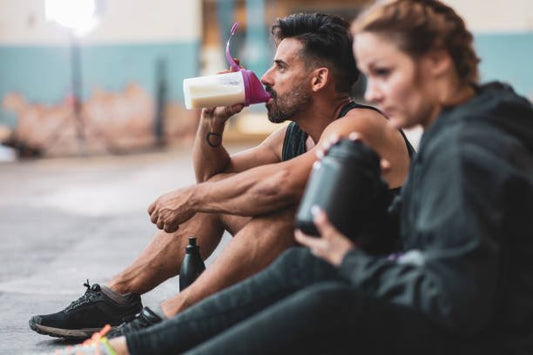 Två personer sitter på golvet i ett gym och dricker proteinshake efter träning.