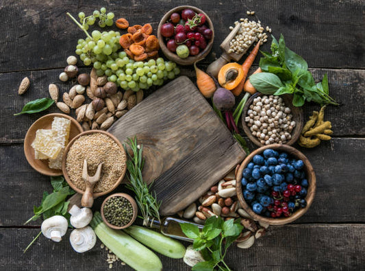 A variety of colorful fruits, nuts, and seeds arranged on a wooden surface with a cutting board in the center.