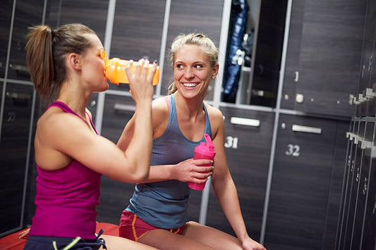 Två kvinnor i omklädningsrum njuter av pre workout-drycker efter träning.
