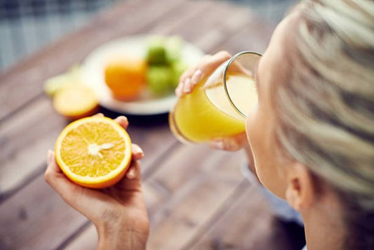 Woman holding an orange half while sipping fresh juice, with fruits on a plate in the background.