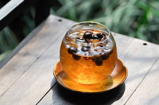 Kopp med färsk cascara-te och svarta bär, placerad på ett träbord i en grönskande miljö.