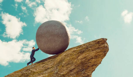 Person pushing a large sphere uphill on a rocky cliff against a bright blue sky.