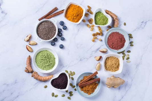 Bowl of various spices and superfoods including turmeric, matcha, and chia seeds arranged artfully on a marble surface.