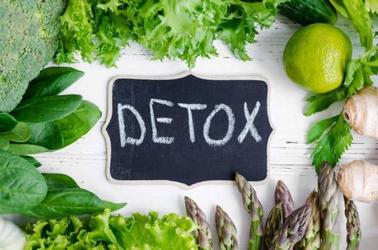 Fresh vegetables and herbs surrounding a chalkboard with the word 'DETOX' written on it.