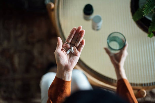Hand holding dietary supplements with a glass of water on a table, representing health and wellness.