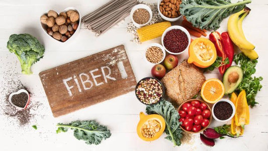 En samling av fiberrika livsmedel som broccoli, nötter, frukter och spannmål, med texten 'FIBER' på en träplatta.