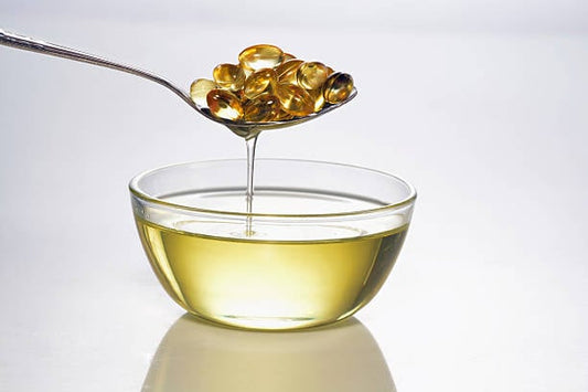 Spoon holding fish oil capsules above a glass bowl of liquid fish oil.