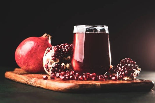 Glas med granatäpplejuice omgiven av granatäpplen på en träskärbräda.
