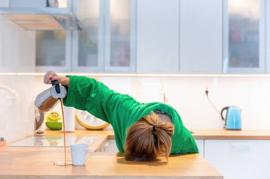Person in a green robe pouring coffee while resting head on kitchen counter, illustrating exhaustion.