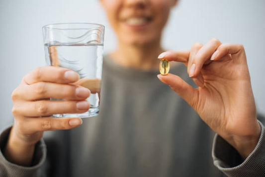 Person holding a glass of water and an omega-3 supplement capsule ready to take.