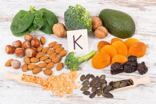Healthy sources of Vitamin K including greens, nuts, and dried fruits arranged with a label.