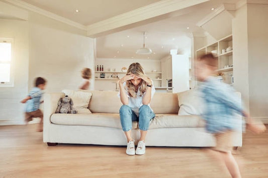 Woman sitting on a couch looking stressed while children play around her in a bright living room.