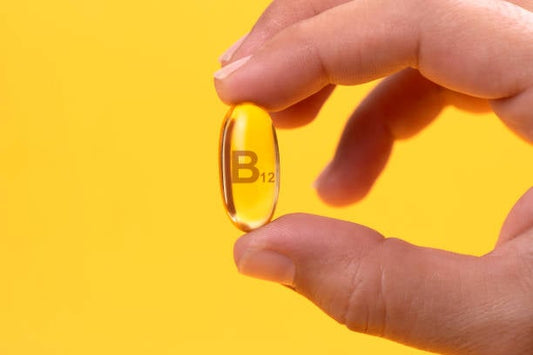 Hand holding a B12 vitamin capsule against a yellow background.