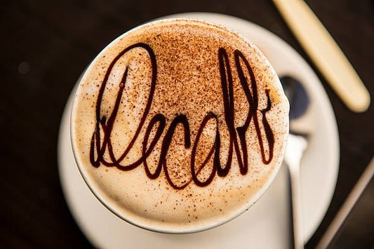 Kopp kaffe med dekoren "decaff" i chokladsås och kanel på toppen, serverad på tallrik.