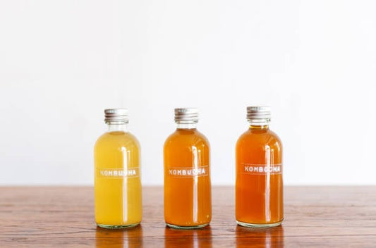 Tre flaskor med kombucha i olika färger på ett träbord.