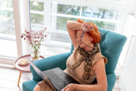 An older woman with red hair sits in a teal chair, looking stressed while working on a laptop.