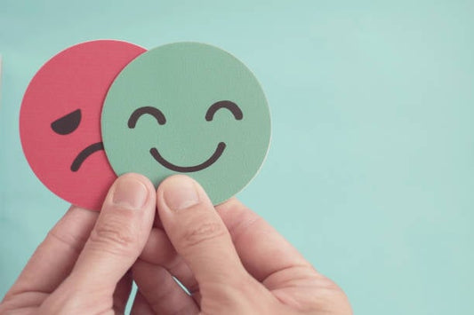 Hands holding green happy face and red sad face cards, symbolizing emotions and well-being.