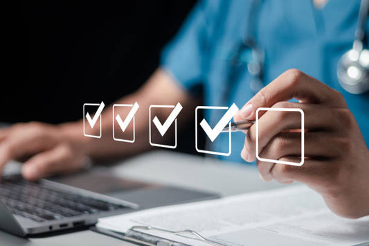 Healthcare professional checking off tasks while using a laptop and stethoscope in a medical environment.