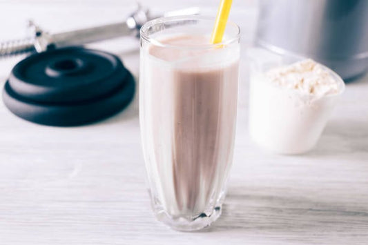Måltidsersättning i glas med sugrör, proteinpulver och vikter i bakgrunden.