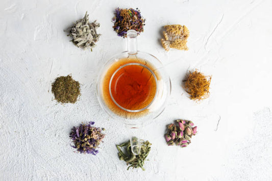 A clear teapot surrounded by various dried herbs, flowers, and spices used to make herbal tea.