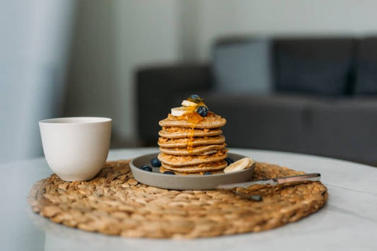 Stack of protein pancakes with banana and blueberries, served on a plate beside a cup of coffee.