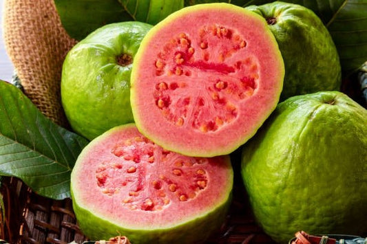 Halved guava revealing pink flesh, surrounded by whole green fruits and leaves, showcasing its vibrant color and texture.