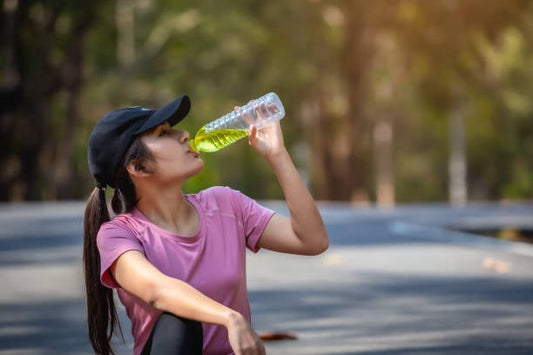 Woman in a pink shirt drinking a green electrolyte beverage outdoors, promoting health and hydration.