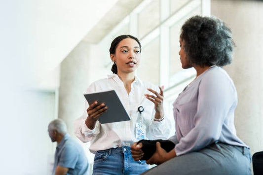 Two women engaged in a discussion, one holding a tablet, in a modern office environment.