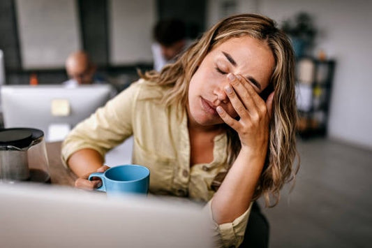 Woman looking tired while holding a blue mug, resting her forehead on her hand in a modern office.